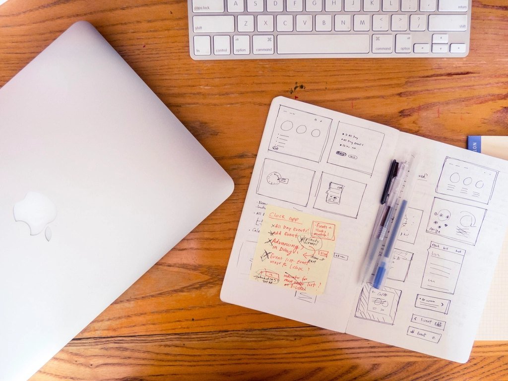 Paper wireframe sketches next to a laptop on a wooden desk